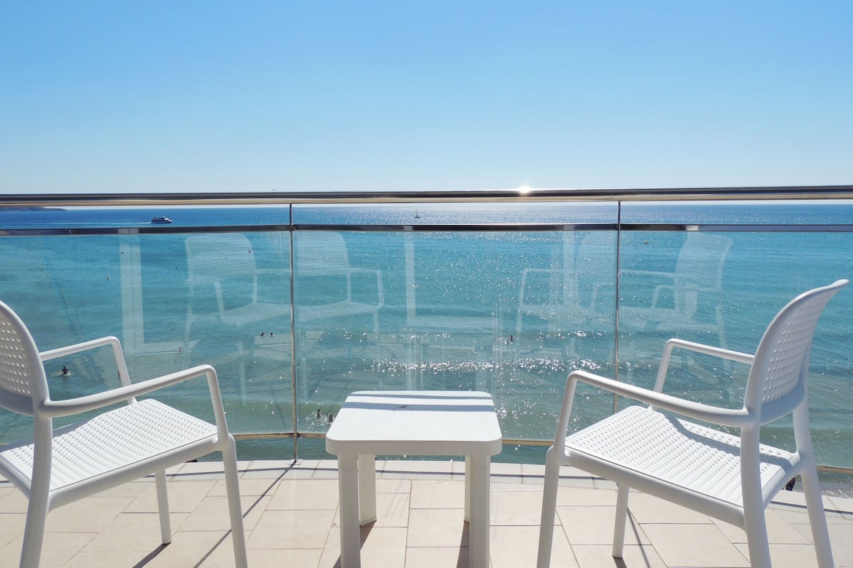Un balcon avec deux chaises blanches et une petite table, d’où l’on peut admirer une mer d’un bleu limpide et une côte lointaine.
