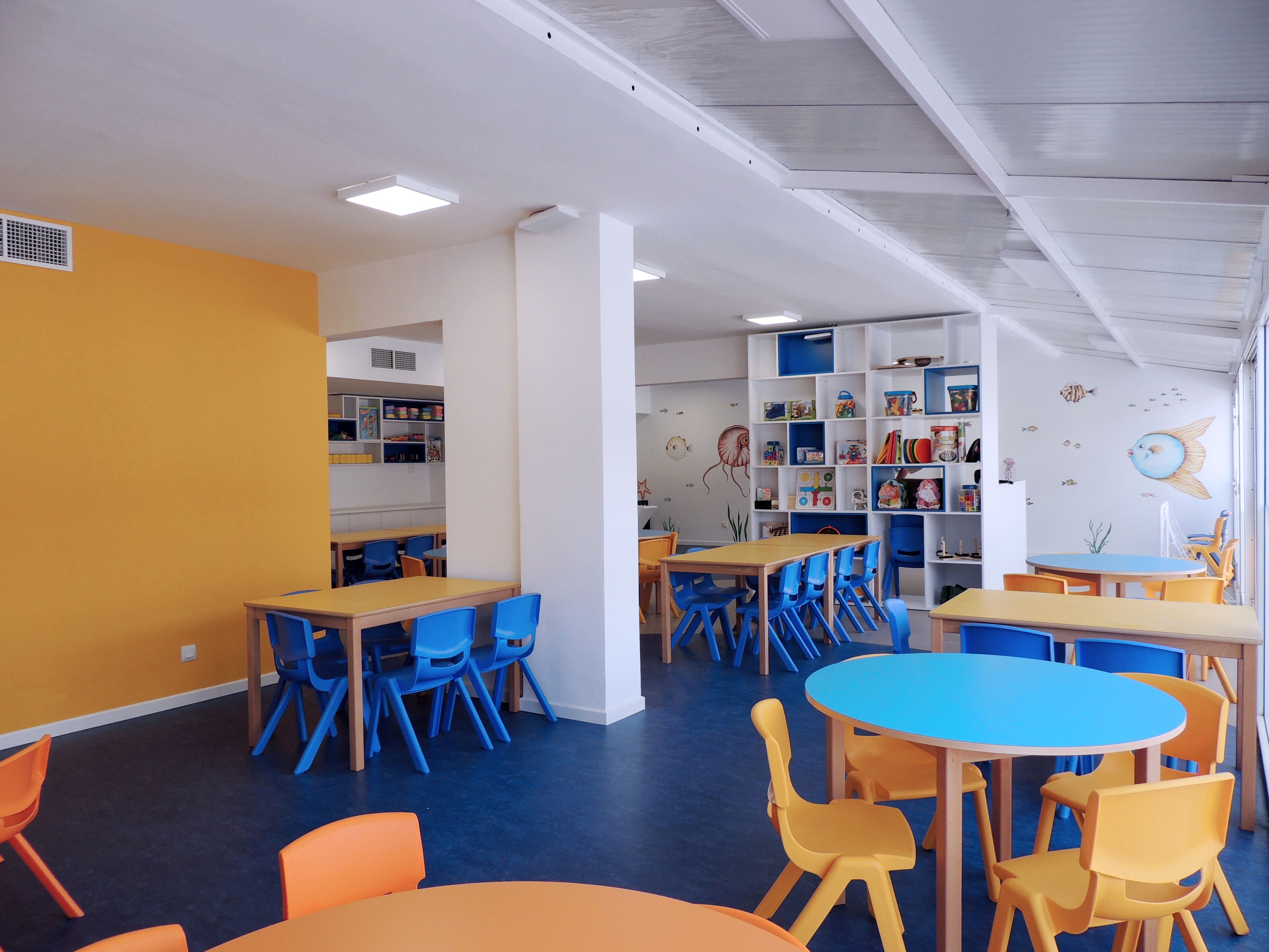 Un cuarto de juegos para niños con mesas y sillas, una estantería y un mural en la pared.