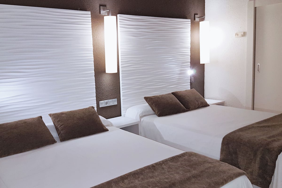 Une chambre d’hôtel avec deux lits, chacun doté de draps blancs et d’oreillers marron. On y trouve des appliques murales et une porte avec une poignée.