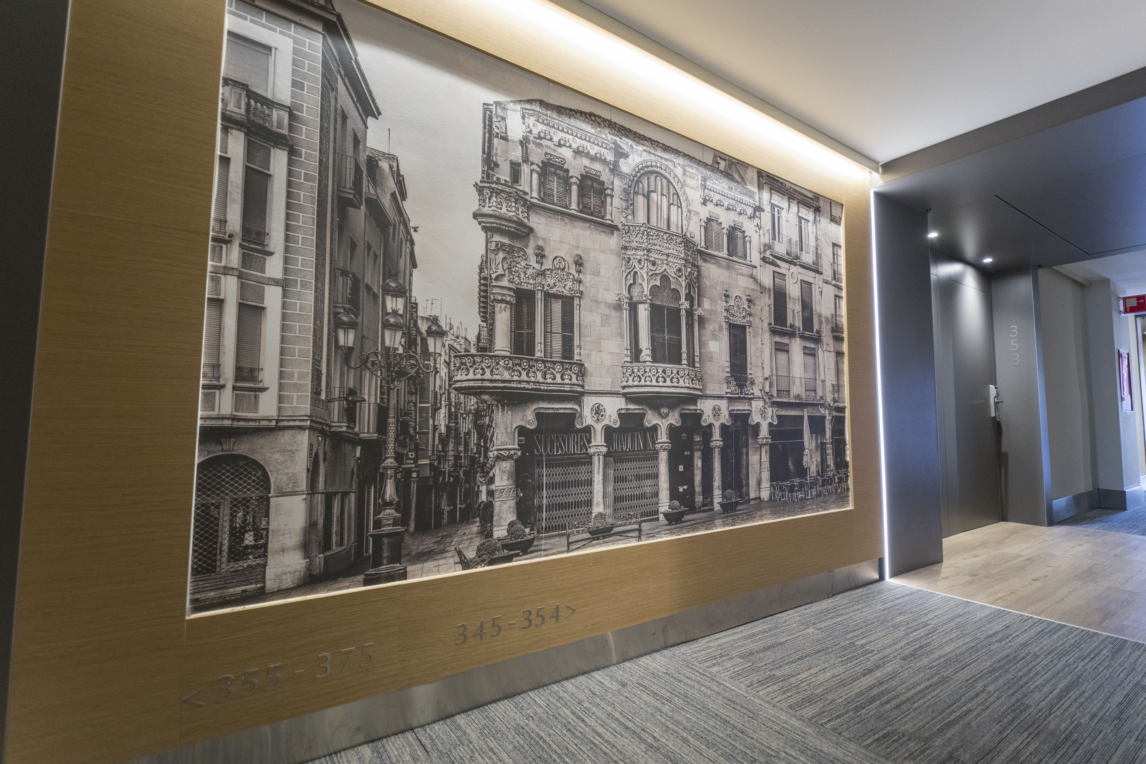 A corridor with a large black and white photograph of historic buildings, a wooden panel with numbers, and a modern elevator.