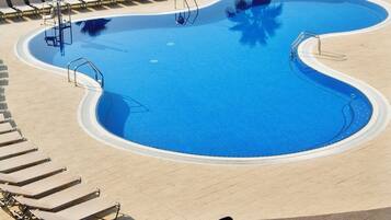 A swimming pool with lounge chairs and tables, a beach in the background, and a palm tree.