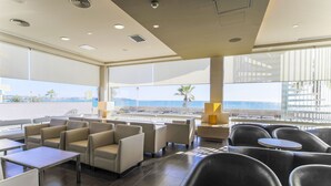 Un área de descanso moderna con asientos y mesas, ventanas amplias con vista al mar y un balcón con barandilla.