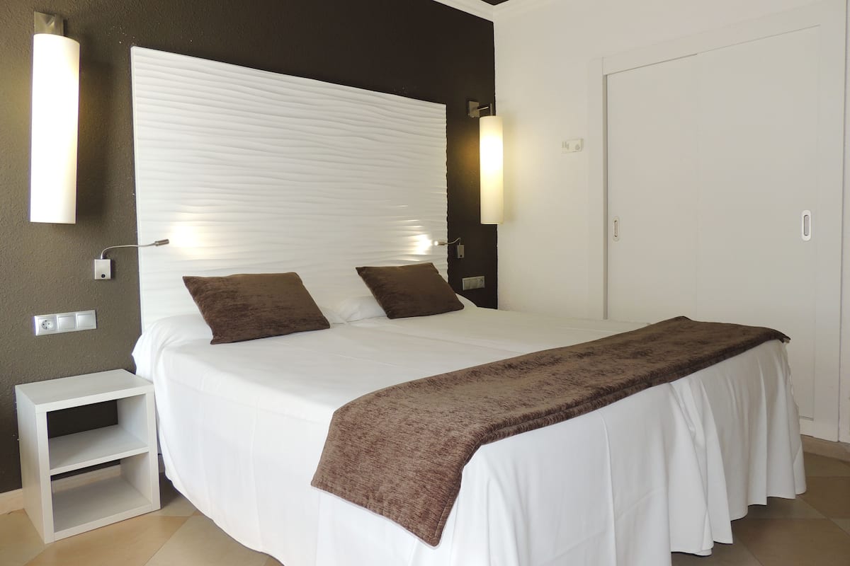 Un lit bien fait, avec une couette blanche et deux oreillers marron, une table de chevet avec une lampe et une armoire avec une porte.