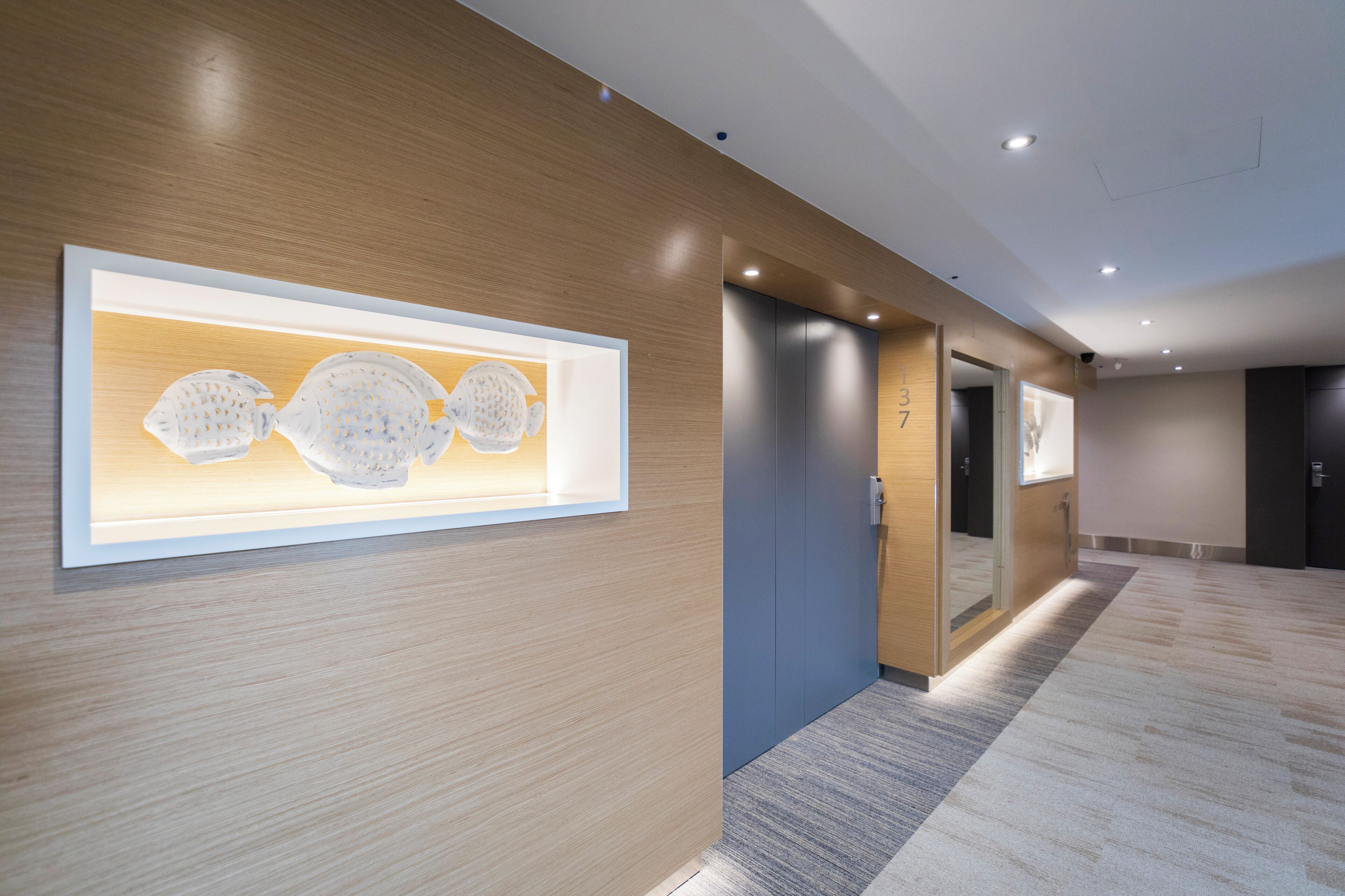 A modern hotel corridor with wooden paneling, recessed lighting, and a framed artwork on the wall.