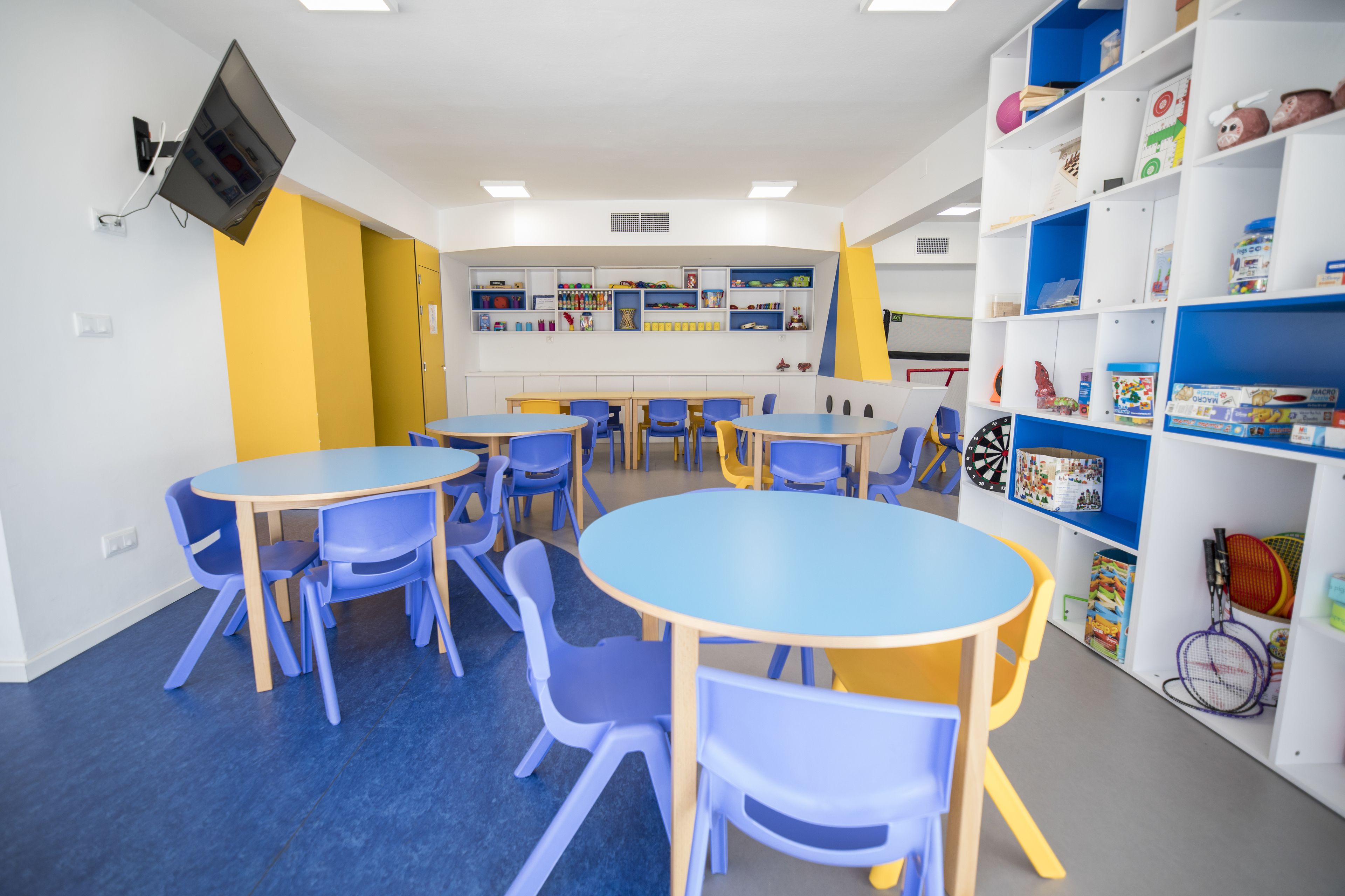 A classroom with blue and yellow tables and chairs, shelves with educational materials, and a mounted television.