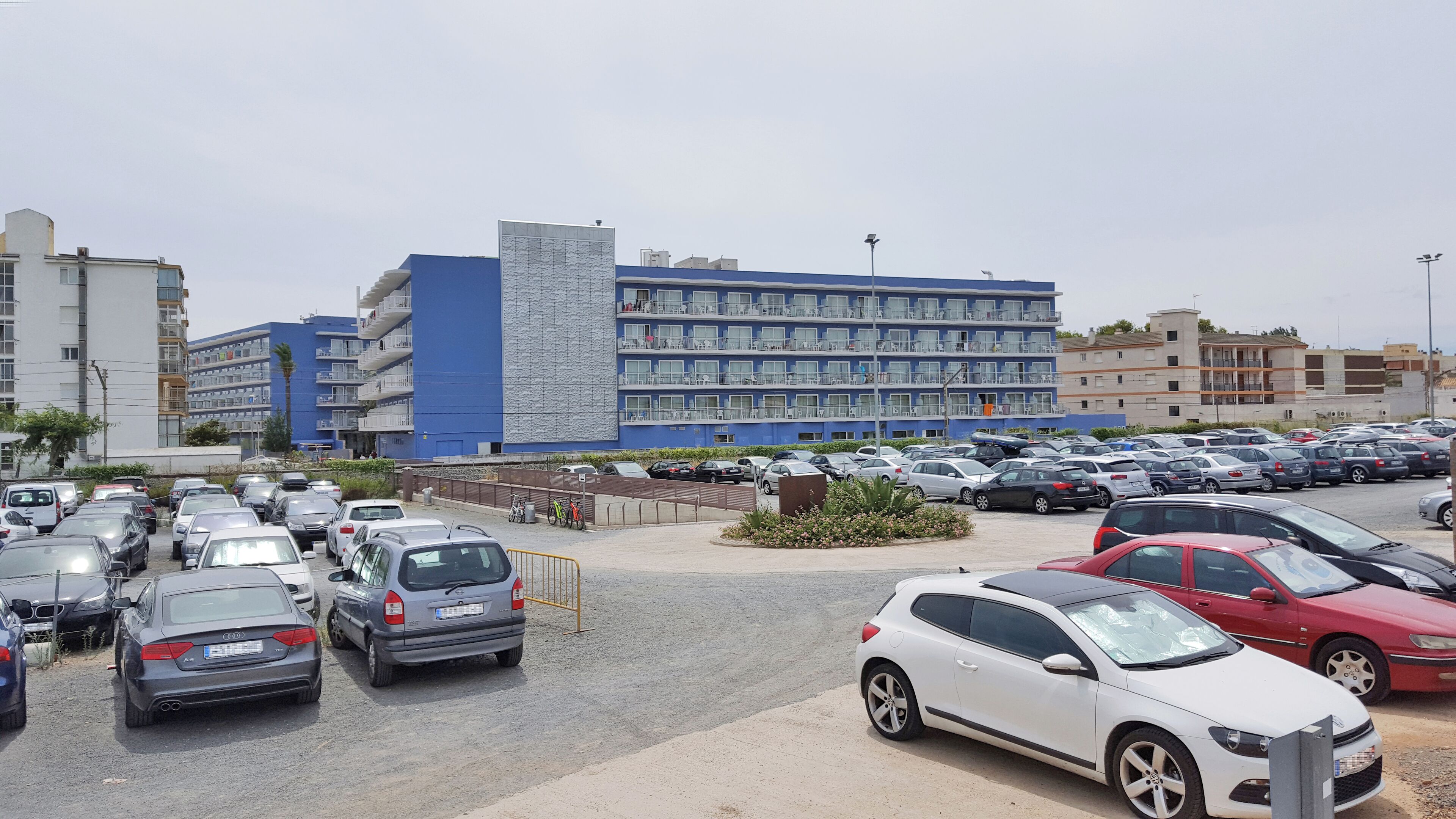 A parking lot with multiple cars, a building with balconies, and a clear sky.