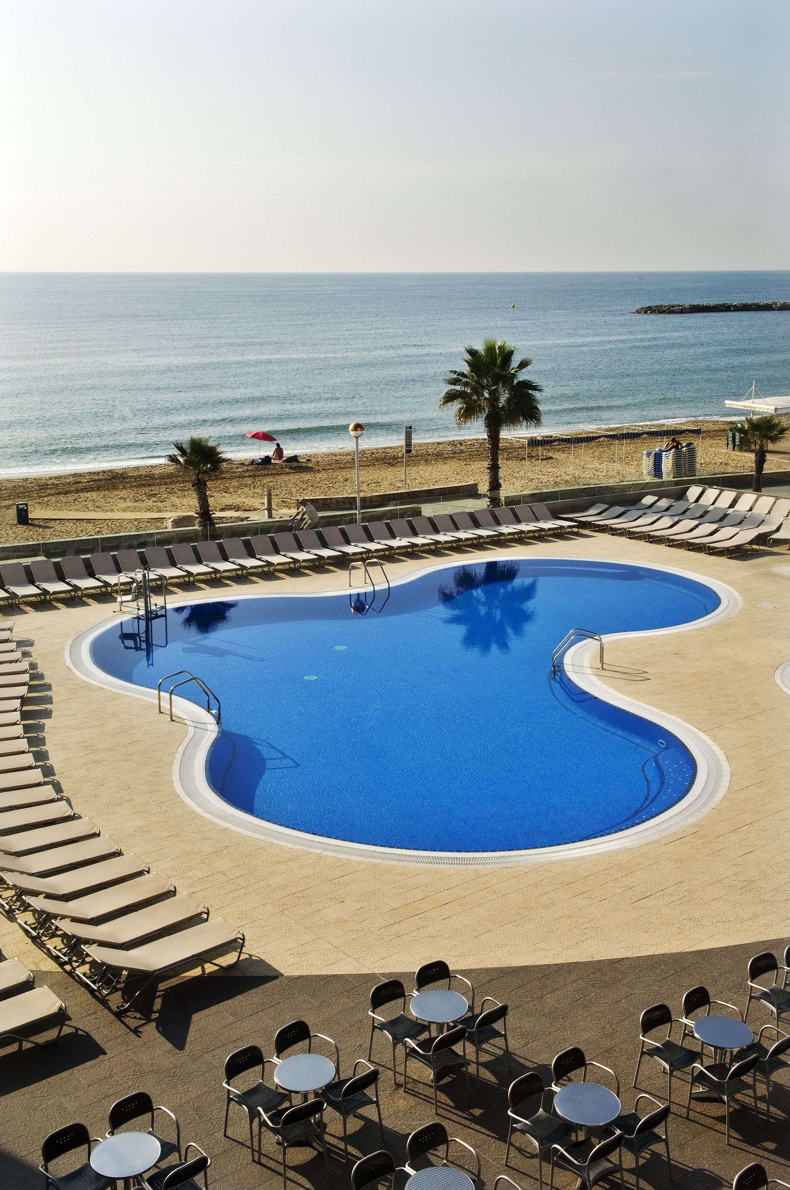 A pool with a curved edge, surrounded by lounge chairs and tables, with a beach and ocean in the background.