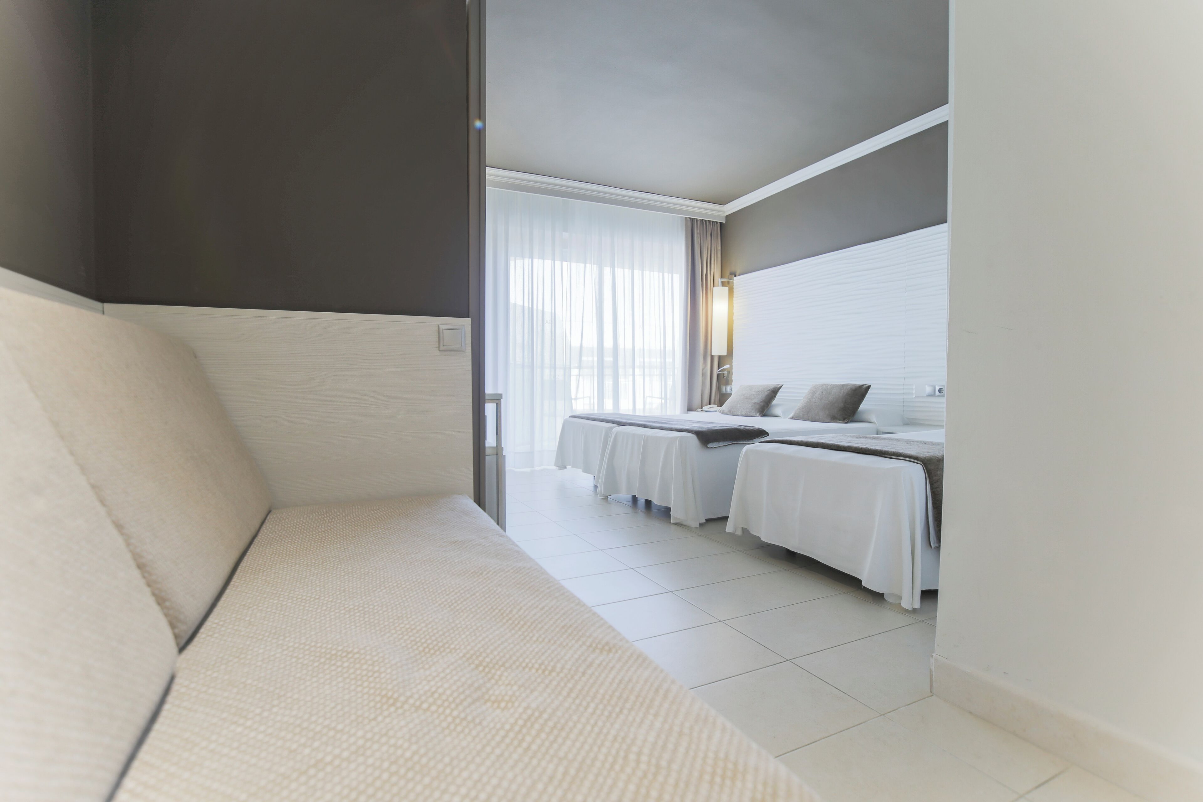 Habitación de hotel con sofá, dos camas con ropa blanca, un ventanal con cortinas translúcidas y una lámpara fijada a la pared.