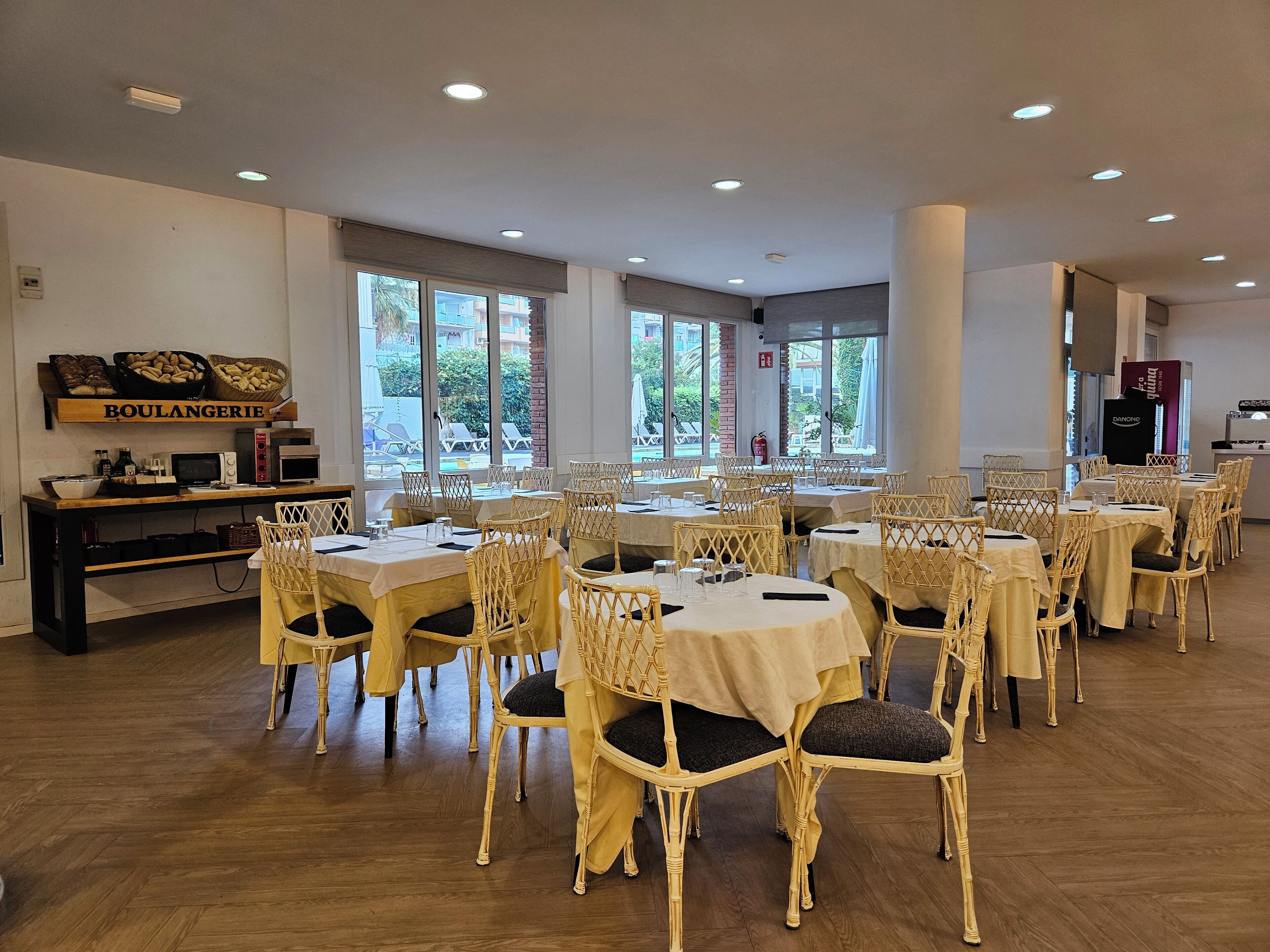 Un restaurante con varias mesas y sillas de mimbre, un letrero que dice "BOULANGERIE" en la pared y grandes ventanales que ofrecen vistas al exterior.