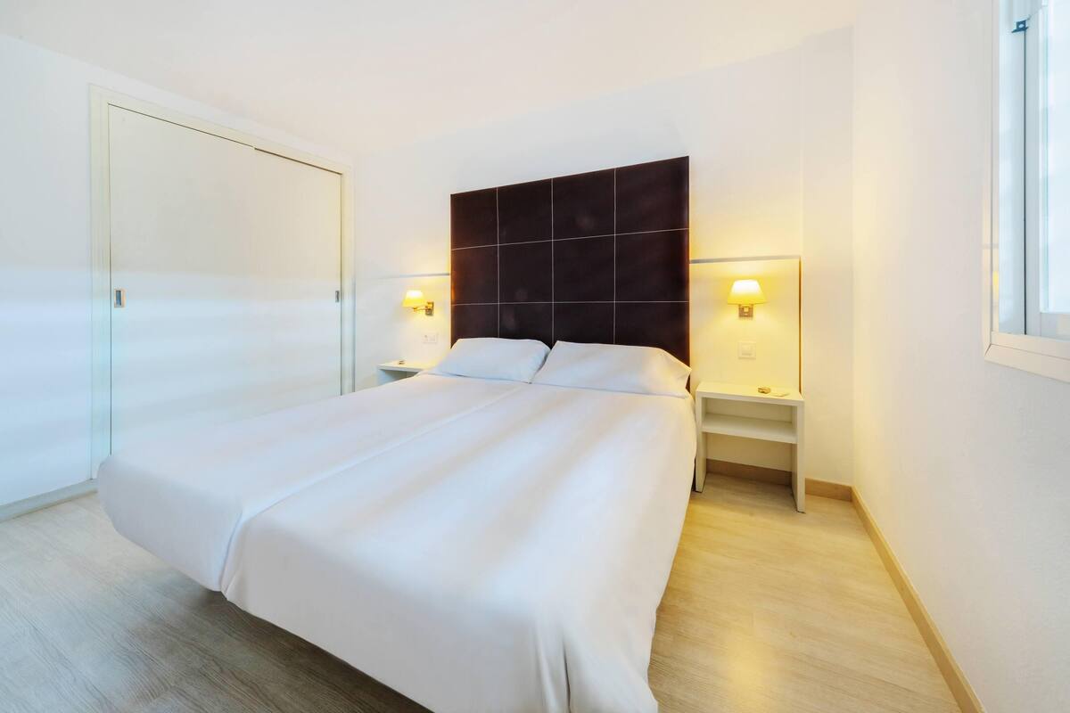 Una cama bien tendida con un cabecero oscuro, dos mesitas de noche con lámparas y suelo de madera.