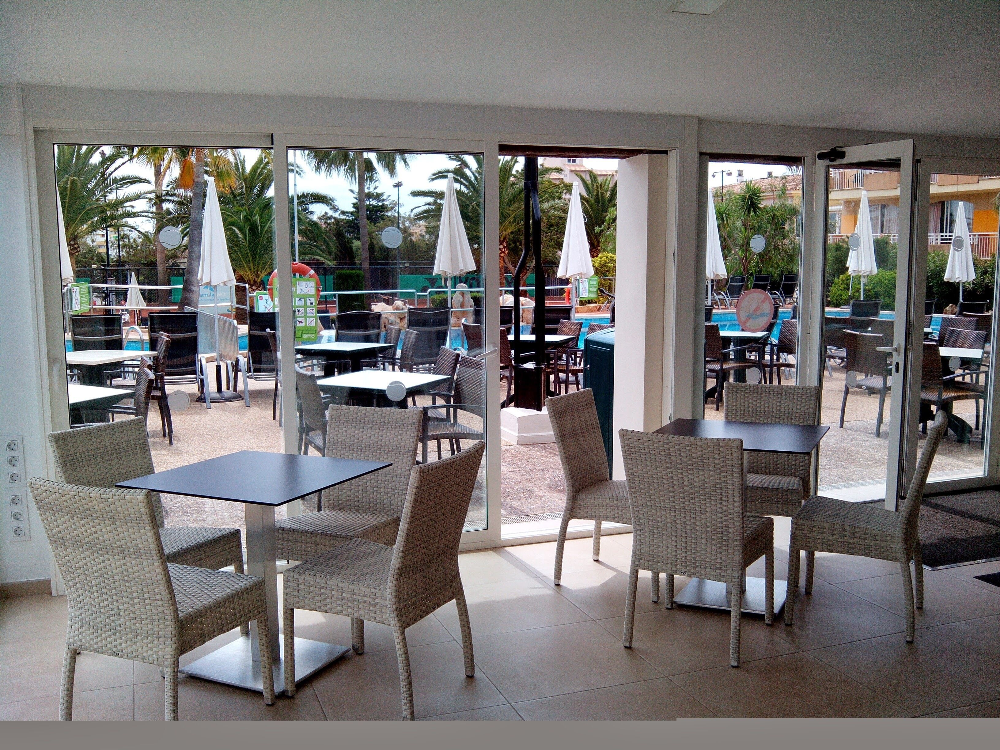 Un espacio de comedor al aire libre con sillas y mesas de mimbre, sombrillas y vistas a una piscina y palmeras.