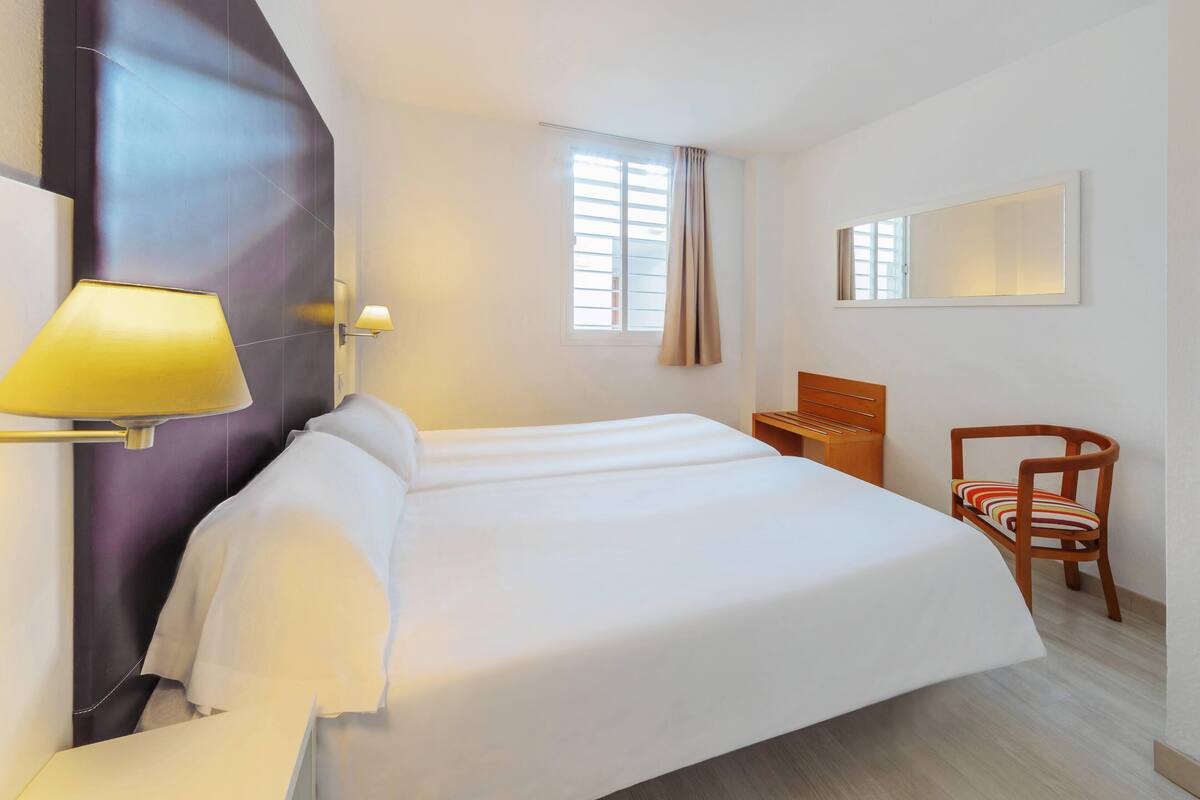 Una habitación de hotel con una cama grande, una mesita de noche de madera, una silla, un espejo montado en la pared y una ventana con cortinas.