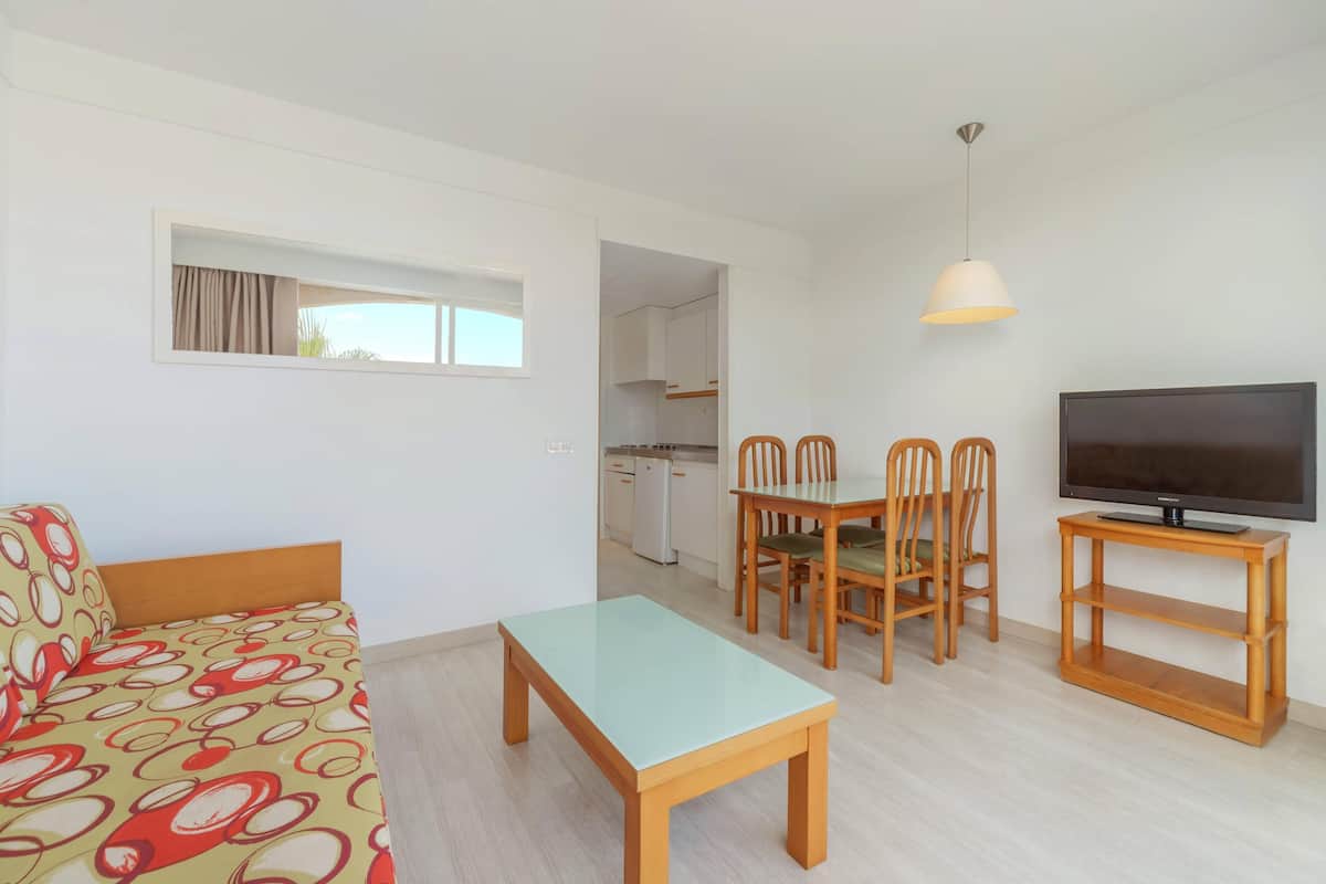 Una sala de estar moderna con un sofá, una mesa de comedor, sillas, un televisor sobre un mueble bajo y una cocina al fondo.