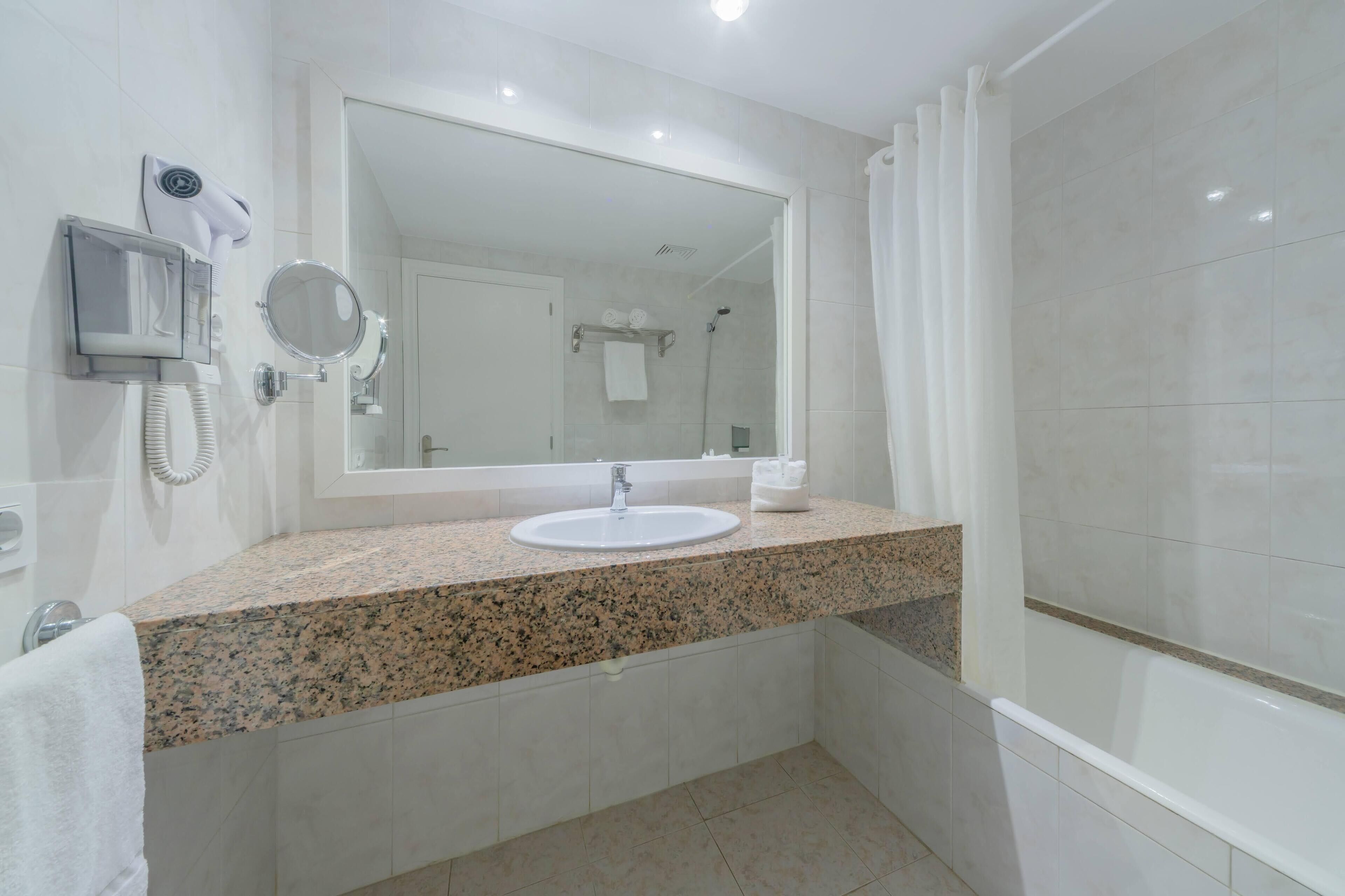 Une salle de bain dotée d’un comptoir en granit, d’un lavabo blanc, d’un grand miroir, d’un sèche-cheveux et d’un espace douche.