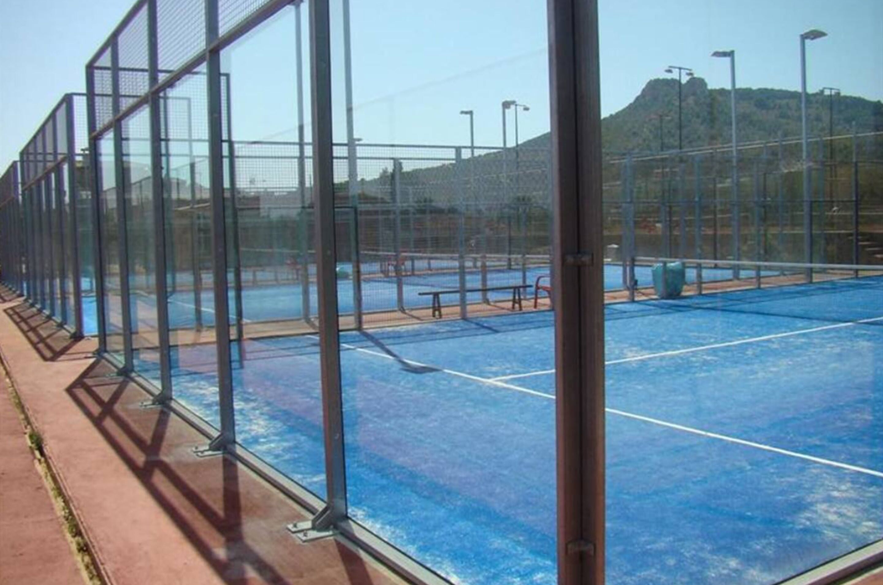 Una cancha de tenis al aire libre con superficie azul, rodeada por una valla de metal, y una montaña al fondo.
