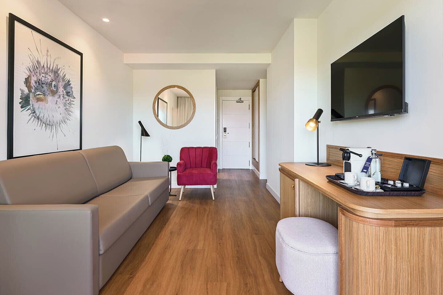 Quarto de hotel moderno com sofá, poltrona vermelha, espelho redondo, escrivaninha com luminária e televisão de tela plana.
