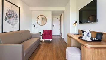 Quarto de hotel moderno com sofá, poltrona vermelha, espelho redondo, escrivaninha com luminária e televisão de tela plana.