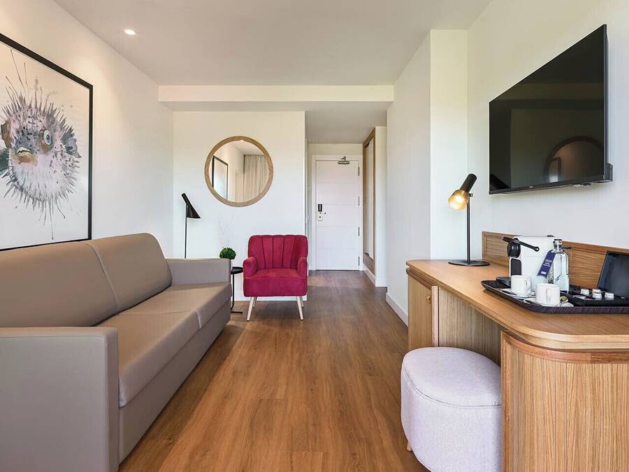 Quarto de hotel moderno com sofá, poltrona vermelha, espelho redondo, escrivaninha com luminária e televisão de tela plana.