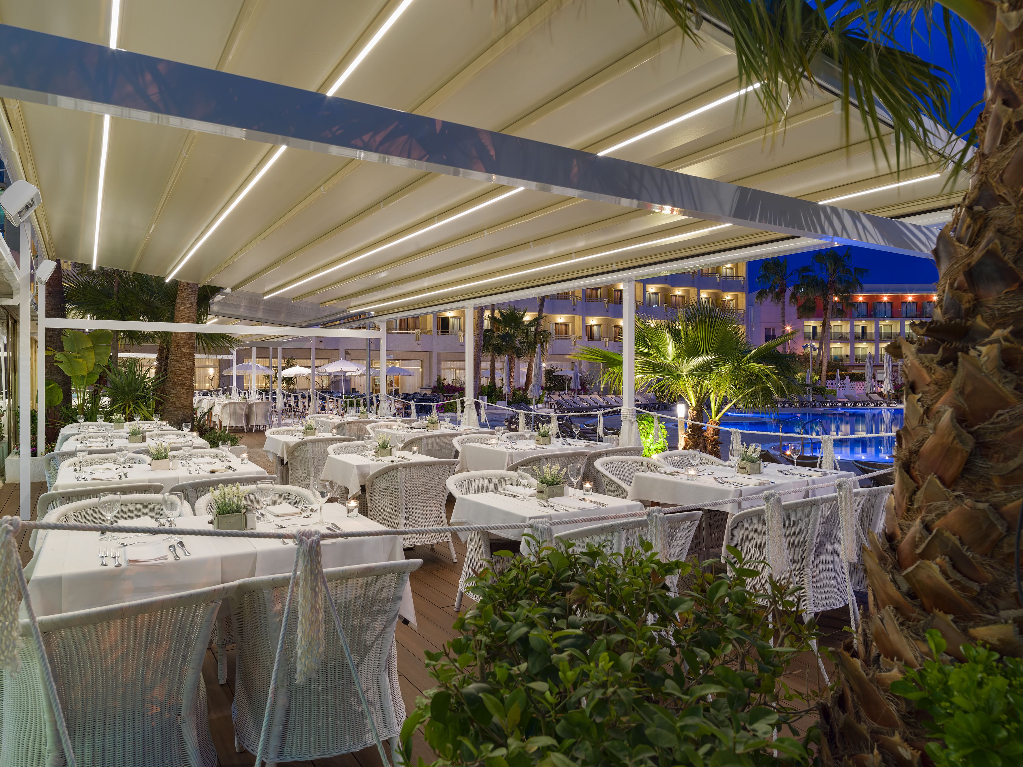 Un espace extérieur aménagé pour les repas, avec des tables et des chaises en osier blanc, une piscine et un bâtiment en arrière-plan.