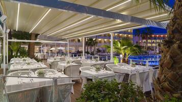 An outdoor dining area with white wicker tables and chairs, a pool, and a building in the background.