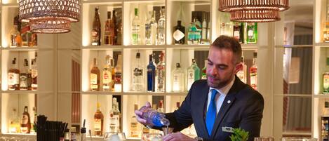 Una zona de bar con un cantinero preparando bebidas, estantes con botellas de licor y lámparas de mimbre colgantes.