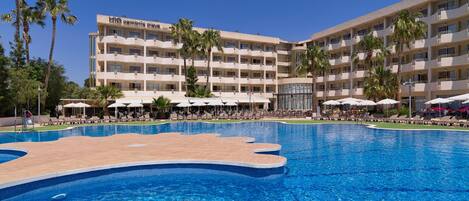A hotel with a swimming pool, sun loungers, and palm trees.