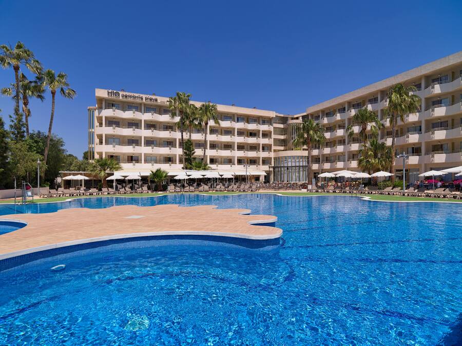 A hotel with a swimming pool, sun loungers, and palm trees.