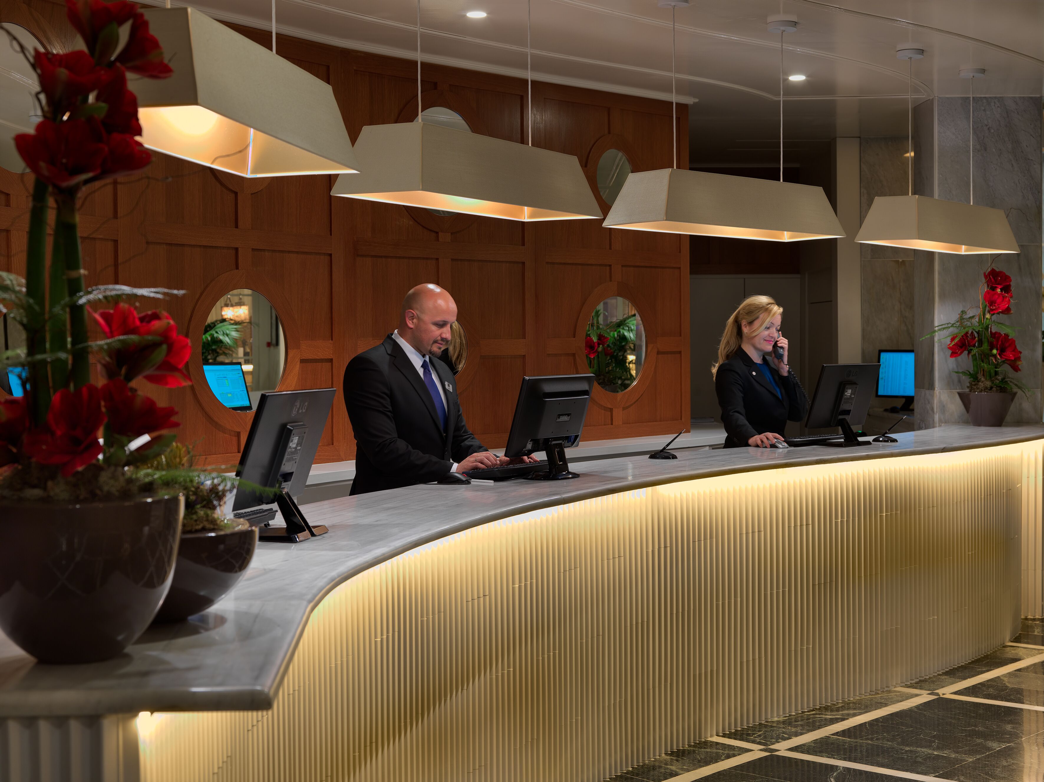 A reception desk with two employees, a wooden panel wall, and a potted plant with red flowers.