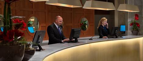 A reception desk with two employees, a wooden panel wall, and a potted plant with red flowers.