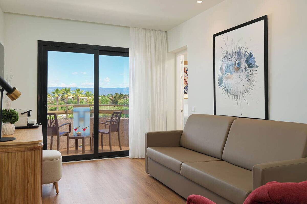 A modern living room with a sofa, a desk with a lamp, a chair, and a large window with a view of palm trees.