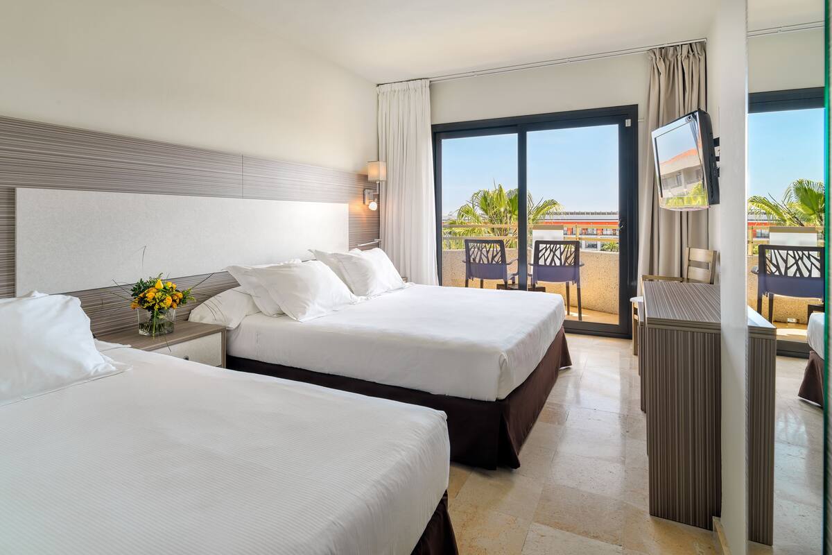A hotel room with two beds, a TV, a balcony with outdoor seating, and a view of palm trees and a building.