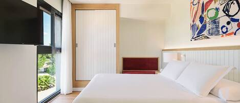 A modern hotel room with a large bed, a ceiling fan, a TV mounted on the wall, and a large window with a view of greenery.