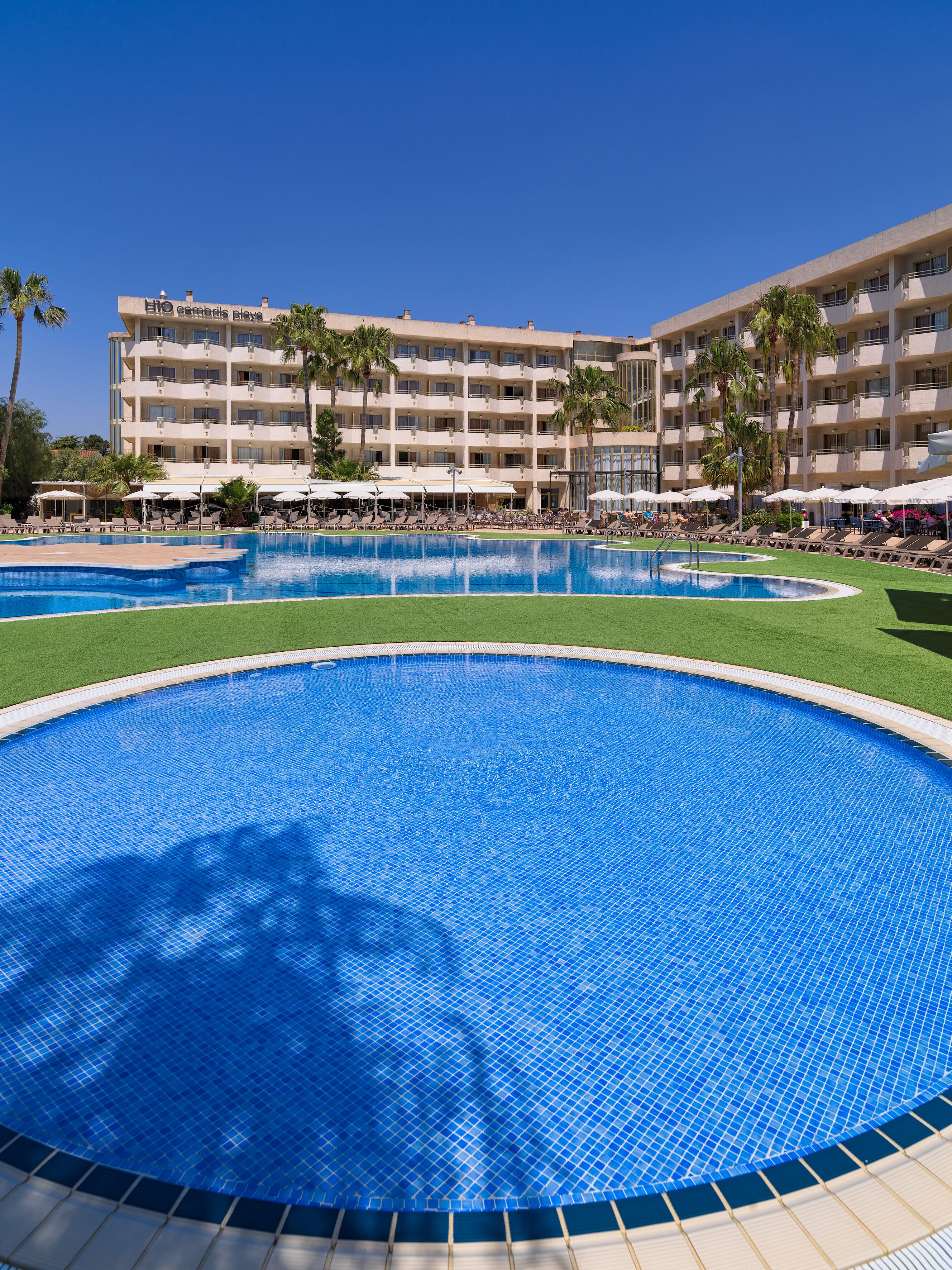 Ein Hotel mit mehreren Balkonen, einem großen zentralen Pool und umliegenden Liegebereichen mit Sonnenschirmen.