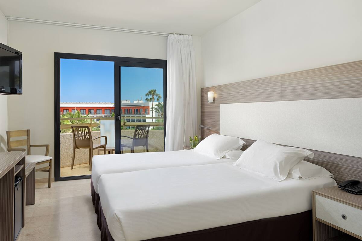 A hotel room with a bed, bedside table, and a desk with a chair. There is a balcony with a view of buildings and palm trees.