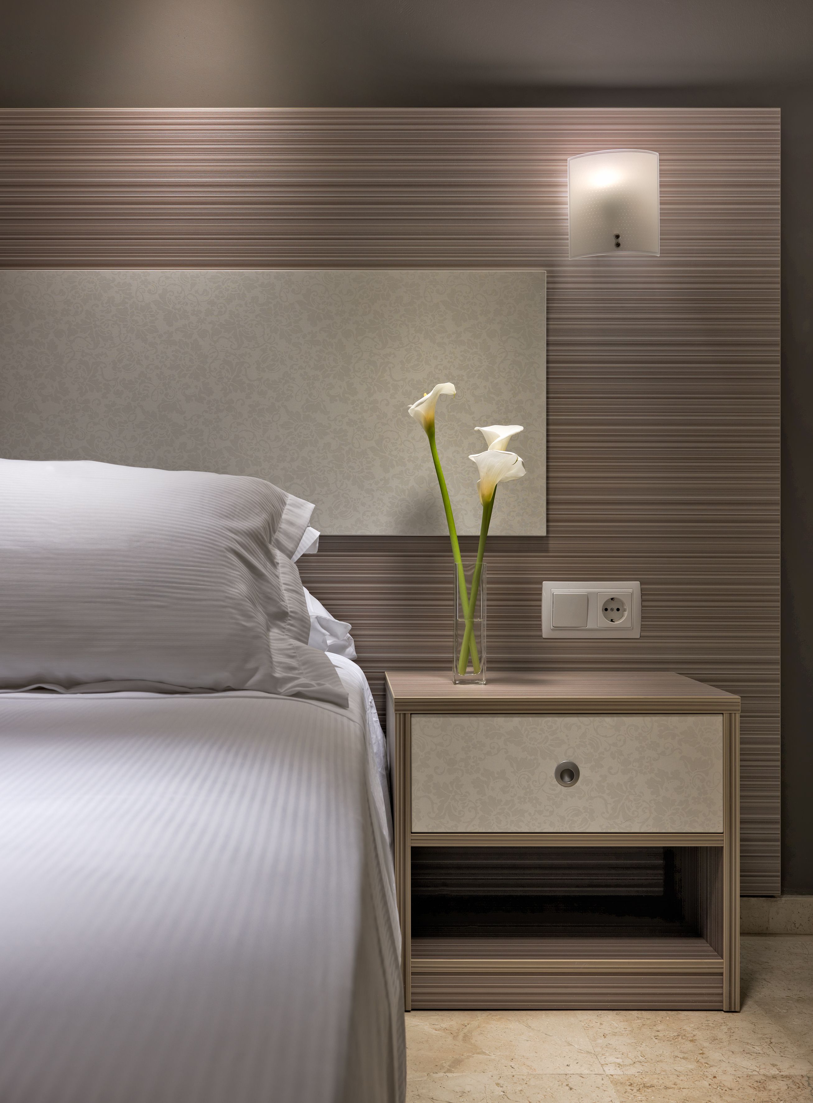 Un dormitorio moderno con cama, una mesita de noche con un jarrón de flores y una lámpara fijada a la pared.