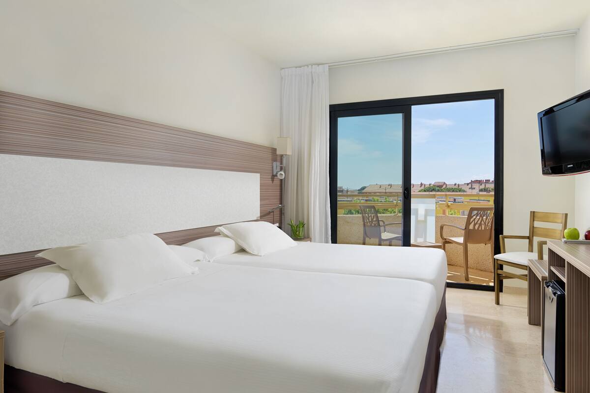A hotel room with two beds, a desk, a chair, a TV, and a balcony with a view.