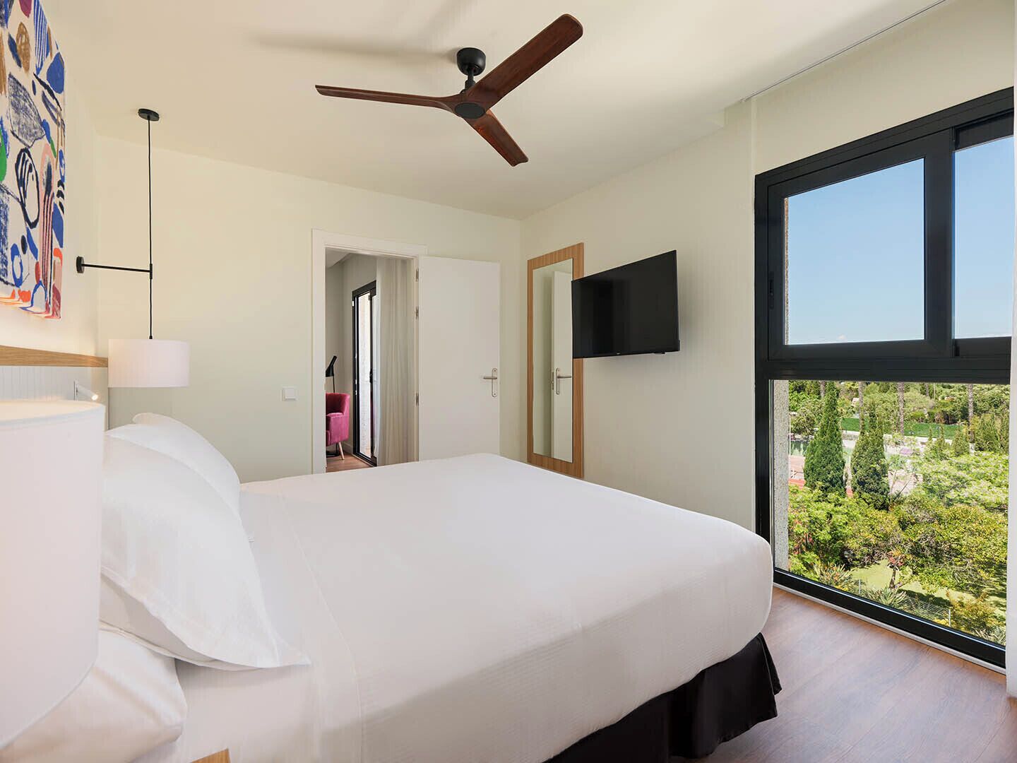 A bedroom with a bed, a ceiling fan, a TV, and a large window with a view.