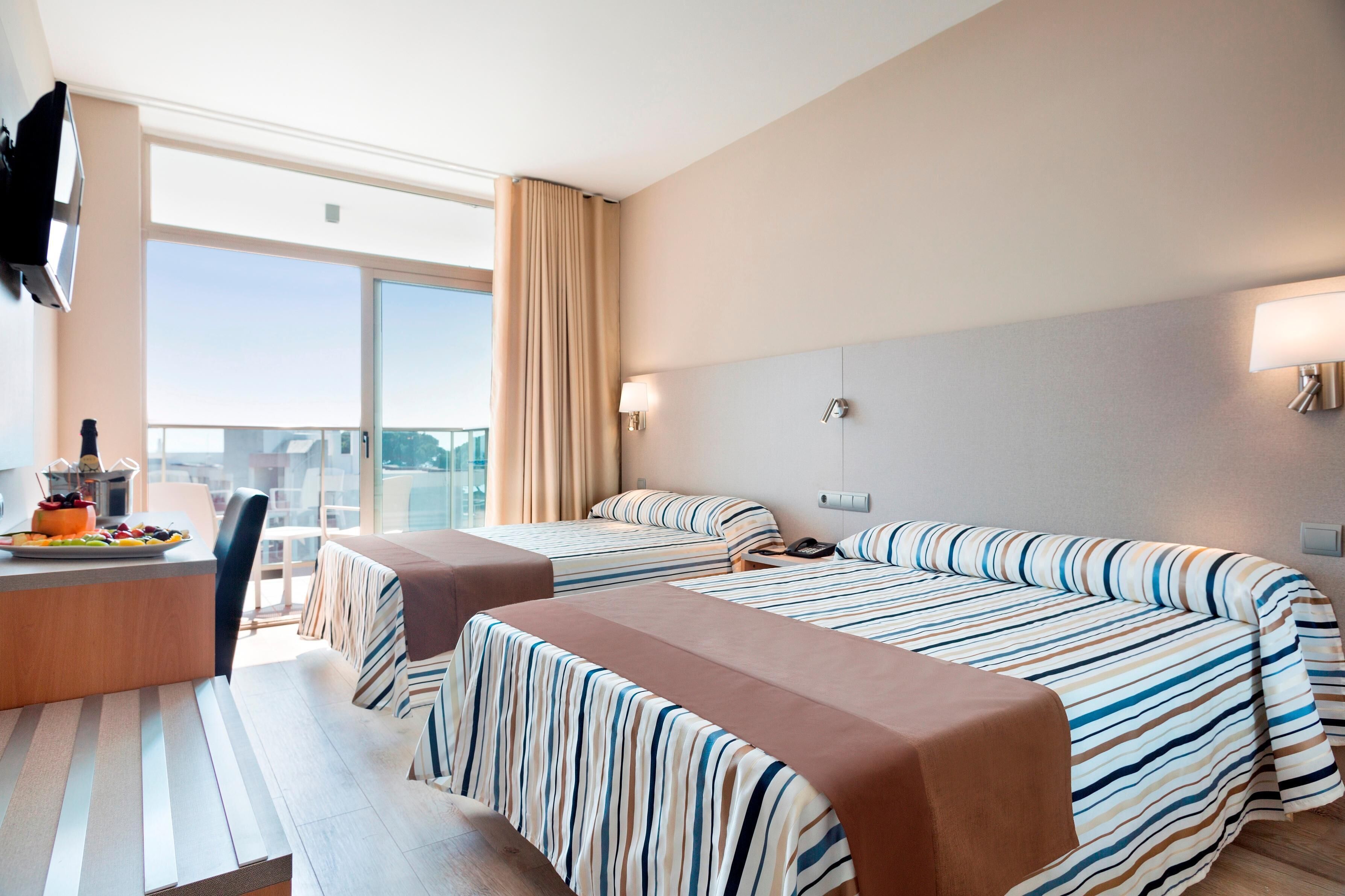 A hotel room with two beds, a desk with a chair, and a large window offering a city view.