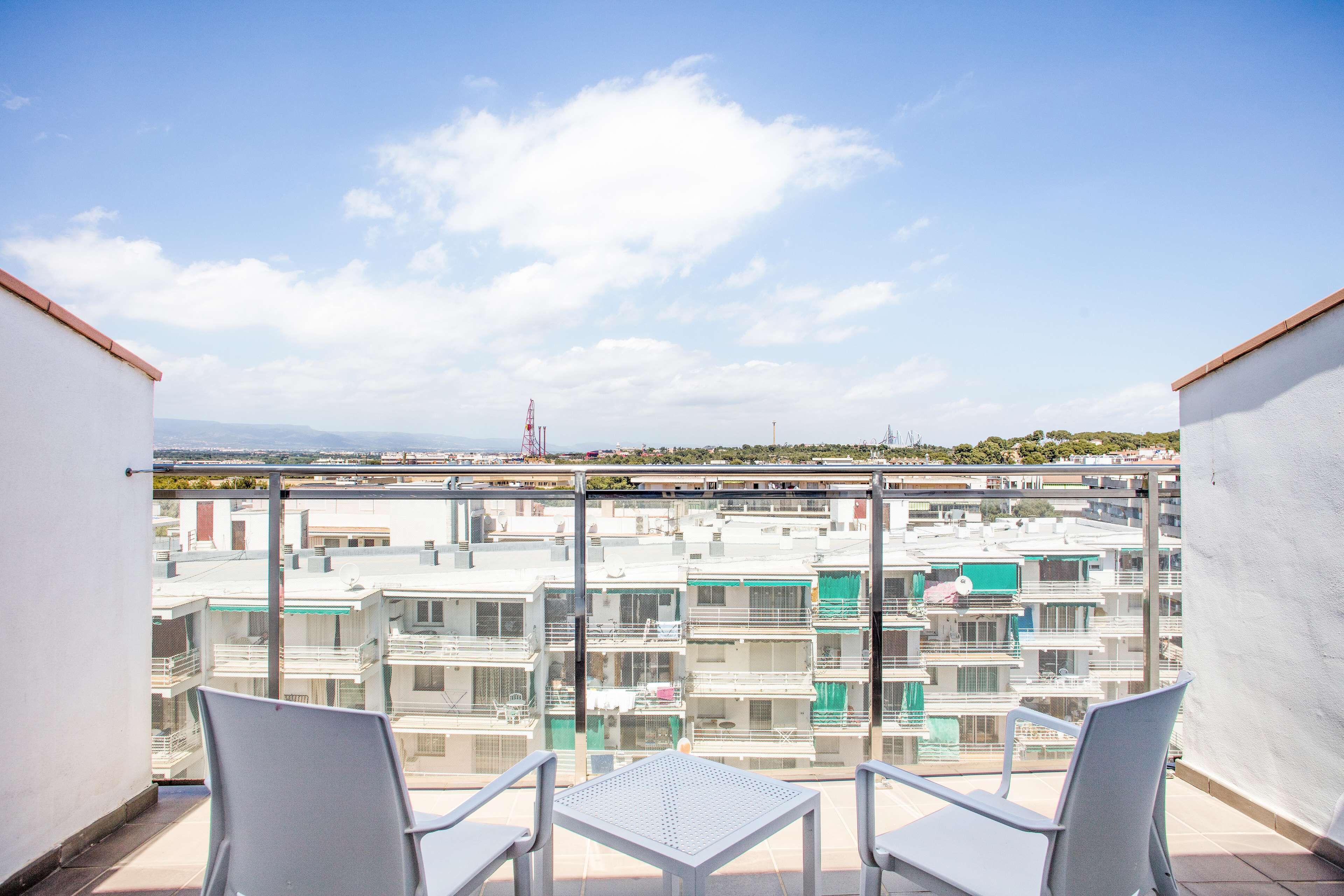 Un balcón con dos sillas y una mesa, con vista a una zona residencial con varios edificios y un cielo despejado.