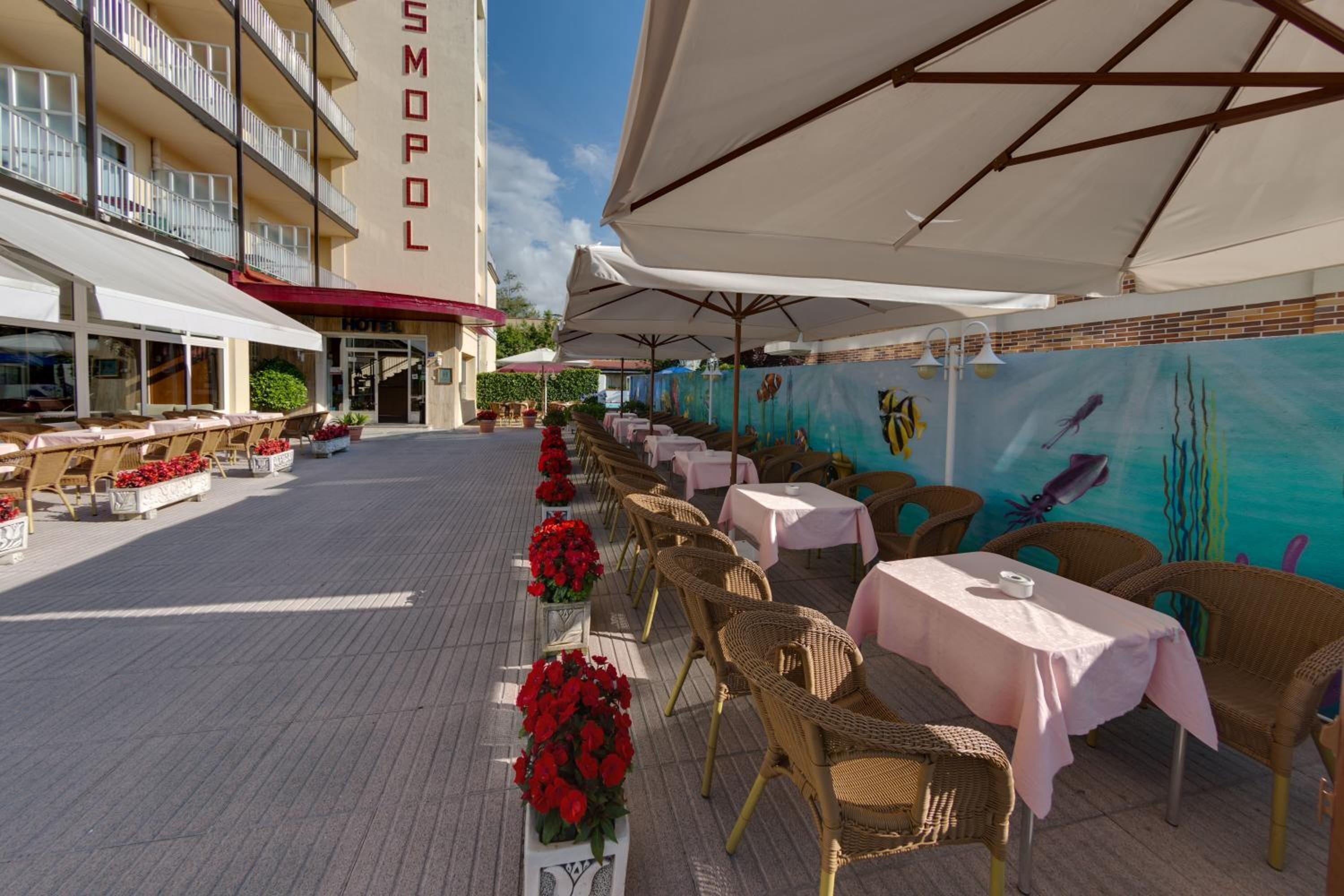 An outdoor dining area with wicker chairs and tables, a mural of marine life, and a hotel building in the background.
