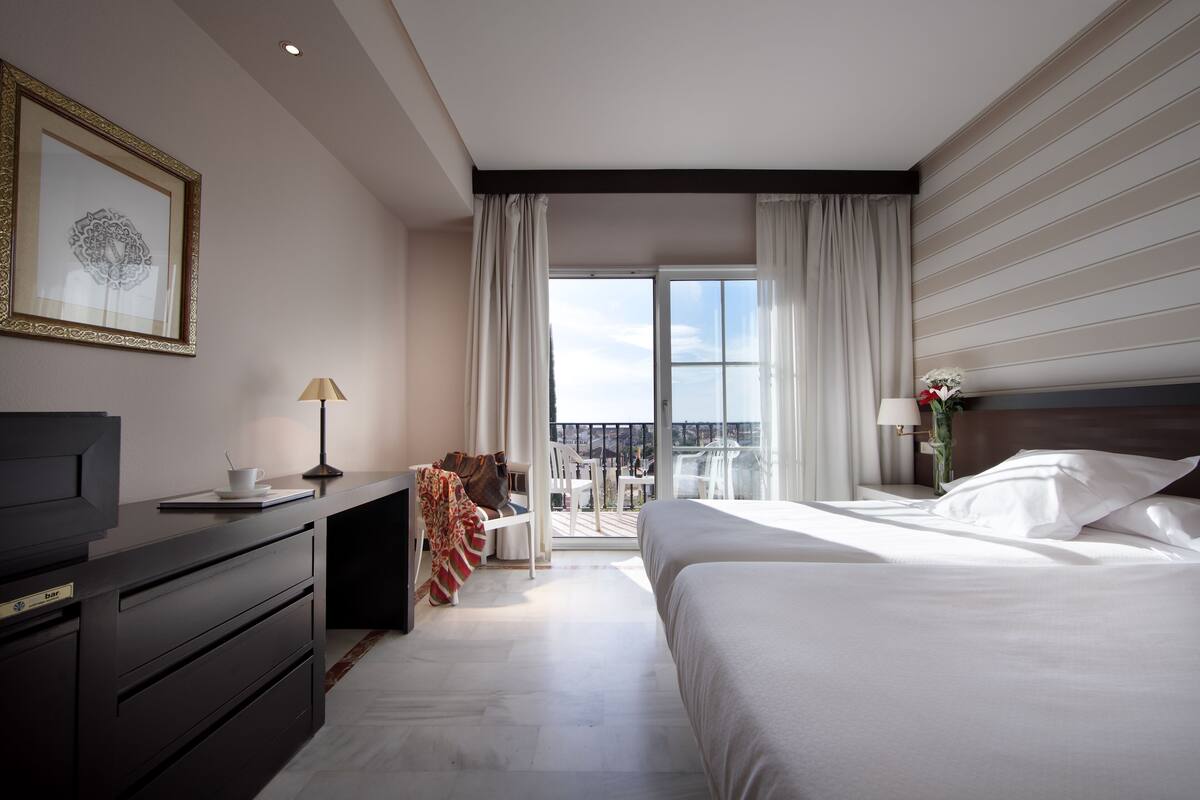 A hotel room with a large bed, a desk with a television, a balcony with a view, and a framed artwork on the wall.