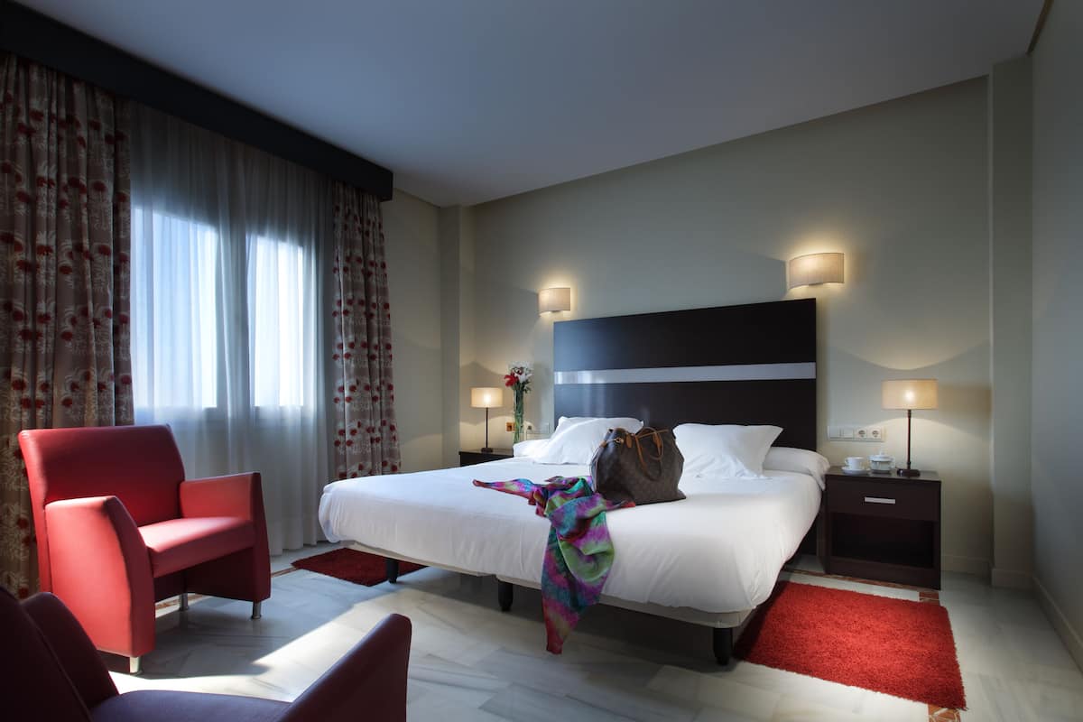 A hotel room with a large bed, a red armchair, a nightstand with a lamp, and a window with curtains.