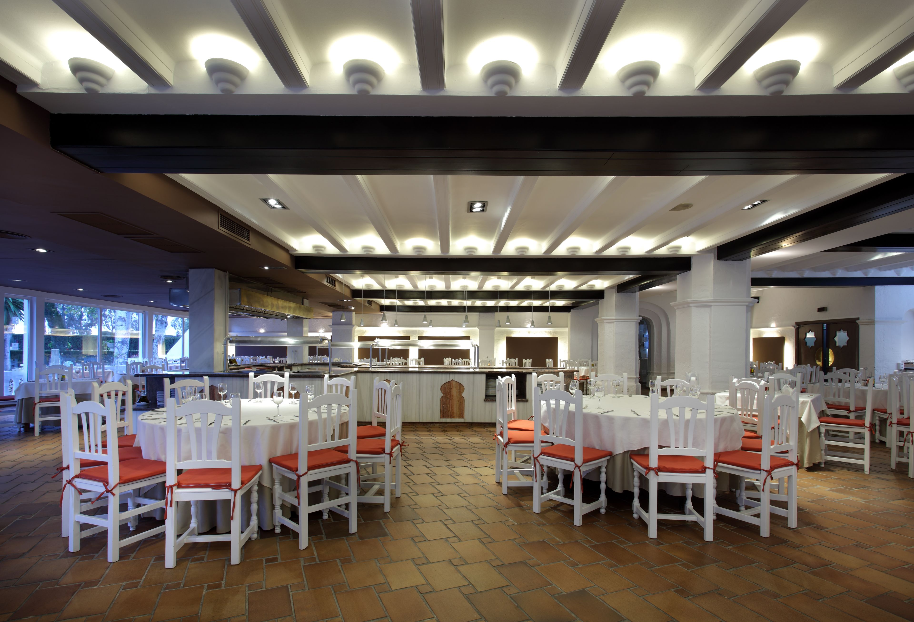 Un vaste espace dédié à la salle à manger, avec des tables et des chaises blanches, un bar central et une vue sur la cuisine.