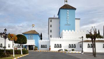 A hotel with a prominent tower labeled "ABADES HOTELES," featuring a white facade with a blue accent, a tiled roof, and a symmetrical entrance with a bench and potted plants.