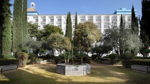 Un patio con un jardín central, rodeado de setos cuidados y árboles altos. Al fondo, un edificio grande con varios balcones y un techo azul característico.