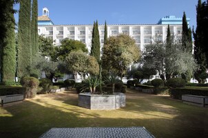Un patio con un jardín central, rodeado de setos cuidados y árboles altos. Al fondo, un edificio grande con varios balcones y un techo azul característico.