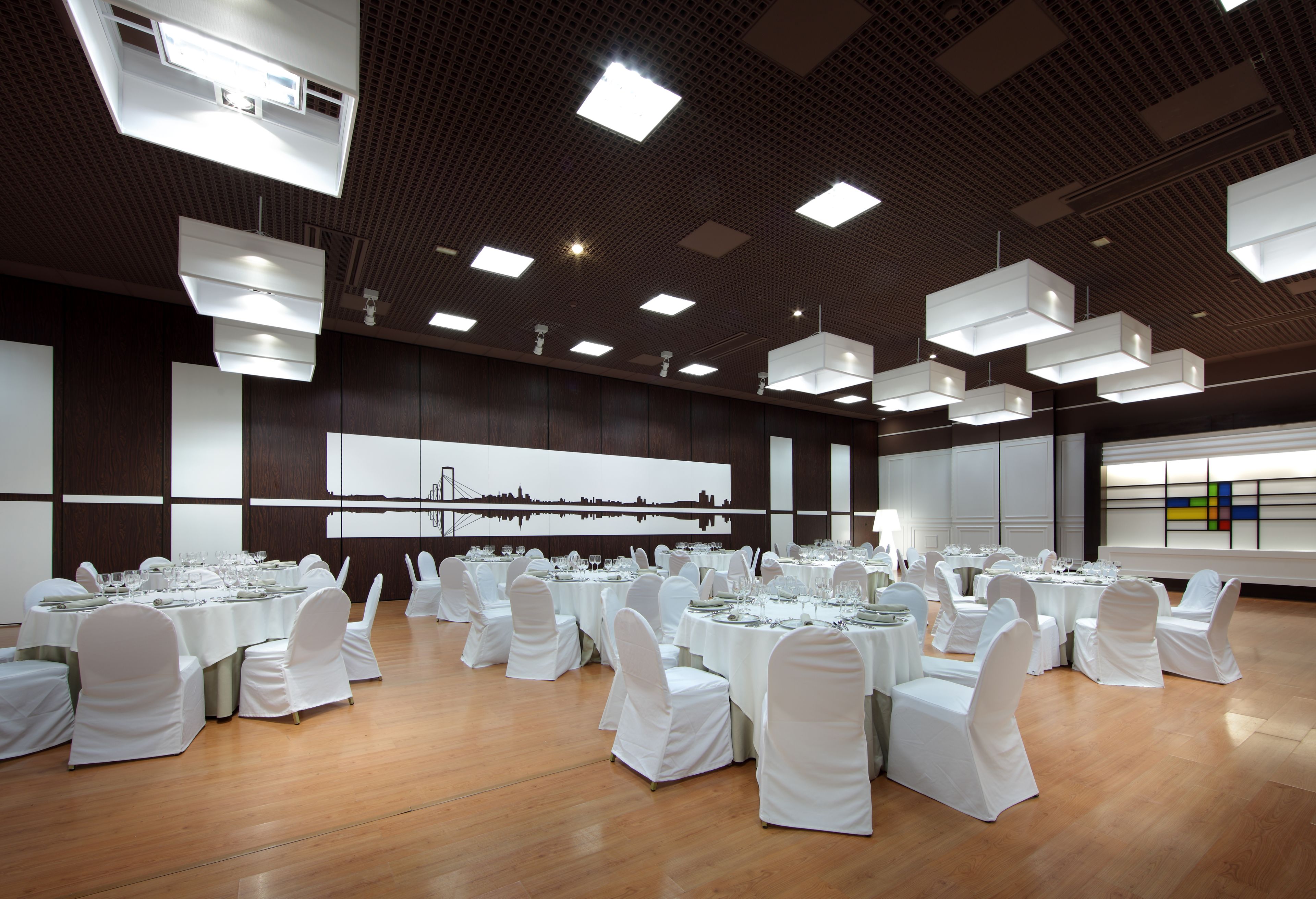 A large banquet hall with multiple round tables set for dining, white chairs, and modern lighting fixtures.