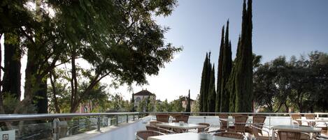 Un patio al aire libre con sillas y mesas de mimbre, un cartel de Bades Hoteles y altos cipreses.