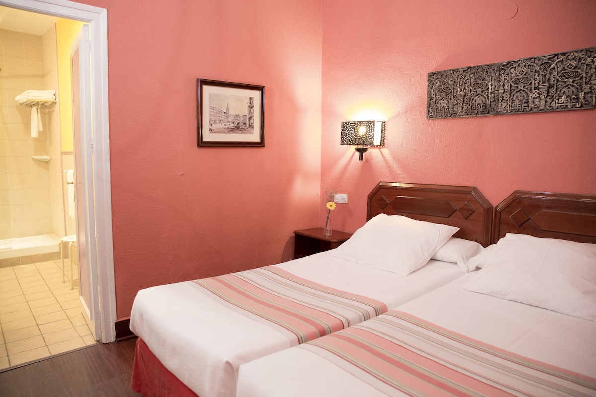 Habitación de hotel con dos camas, un cuadro enmarcado en la pared, una mesita de noche con lámpara y una zona de ducha visible a través de una puerta abierta.