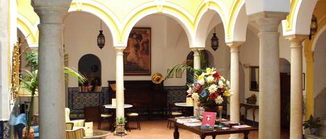 A spacious lobby with a high ceiling, featuring a central table with a vase of flowers, surrounded by seating areas and columns.